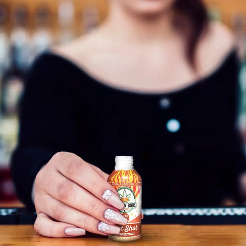 A woman with manicured nails and a black off-the-shoulder top holds an open bottle of Drinkin’ Buds Hot Shot Cinnamon in her hand across a wooden bar surface.