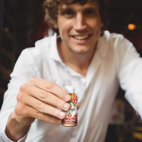 A smiling man in a white shirt holds an open bottle of Drinkin’ Buds Hot Shot Cinnamon toward the camera, suggesting a friendly or social setting.