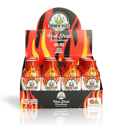 A retail display of Drinkin’ Buds Hot Shot Cinnamon shots, with nine small red bottles featuring flame designs arranged in a black and red tray. The label advertises 10mg THC and 10mg CBD per shot and includes a 21+ warning.
