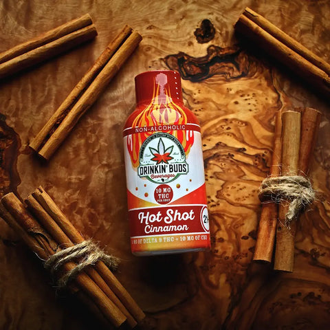 A bottle of Drinkin’ Buds Hot Shot Cinnamon lies on a wooden surface surrounded by bundles of cinnamon sticks, emphasizing the product’s flavor theme in a rustic, natural setting.