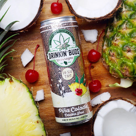A chilled can of Drinkin' Buds Piña Colada Double Shot is surrounded by fresh pineapples, coconuts, and cherries on a wooden surface.