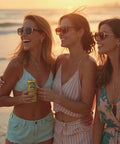 Three smiling women in summer outfits and sunglasses stand together on a beach at sunset, with one holding a yellow can of Drinkin' Buds Spiked Lemonade.