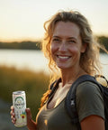 A smiling woman with a backpack holds a can of Drinkin’ Buds Piña Colada beverage outdoors near a lake at sunset.