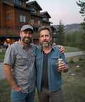 Two men pose arm-in-arm outdoors near a lodge, with one holding a Drinkin’ Buds beverage can.
