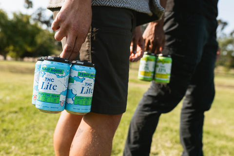 Person holding cans of 'THC Lite' brew in a park setting