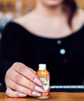 A woman with manicured nails and a black off-the-shoulder top holds an open bottle of Drinkin’ Buds Hot Shot Cinnamon in her hand across a wooden bar surface.