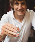 A smiling man in a white shirt holds an open bottle of Drinkin’ Buds Hot Shot Cinnamon toward the camera, suggesting a friendly or social setting.