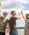 Three men wearing sunglasses raise cans of Drinkin’ Buds Margarita Double Shot in celebration by a waterfront under a partly cloudy sky.