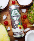 A chilled can of Drinkin' Buds Piña Colada Double Shot is surrounded by fresh pineapples, coconuts, and cherries on a wooden surface.
