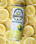 The image features a can of "Drinkin' Buds Beverages" in Spiked Lemonade flavor, placed on a background of fresh lemon slices. The can design highlights a cannabis leaf, the slogan "Taste the New Buzz," and a depiction of a lemonade glass with a straw, emphasizing a cool and citrusy experience infused with 5 mg of Delta 9 THC and 5 mg of CBD.