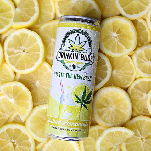 The image features a can of "Drinkin' Buds Beverages" in Spiked Lemonade flavor, placed on a background of fresh lemon slices. The can design highlights a cannabis leaf, the slogan "Taste the New Buzz," and a depiction of a lemonade glass with a straw, emphasizing a cool and citrusy experience infused with 5 mg of Delta 9 THC and 5 mg of CBD.