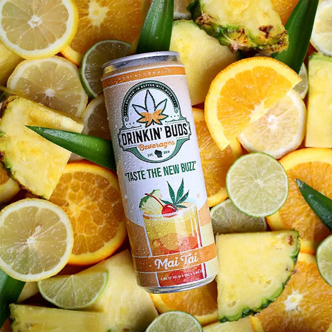 The image showcases a "Drinkin' Buds Beverages" Mai Tai can, surrounded by vibrant slices of pineapple, orange, lime, and lemon. The tropical arrangement highlights the drink's fruity, THC- and CBD-infused essence.