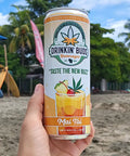 The image shows a hand holding a "Drinkin' Buds Beverages" Mai Tai can on a sunny beach, with surfboards and palm trees in the background. The scene evokes a tropical, relaxed vibe, perfect for enjoying the fruity, infused drink.
