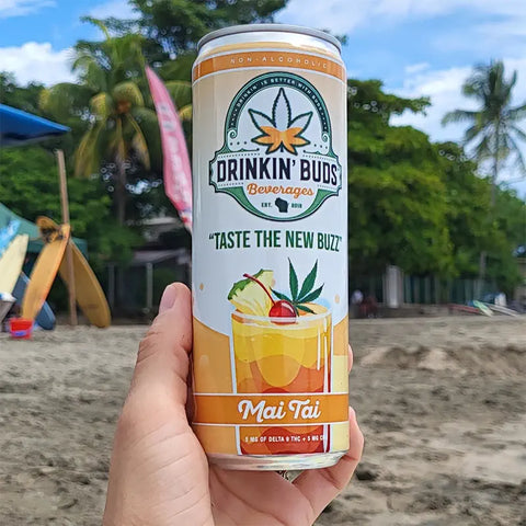 The image shows a hand holding a "Drinkin' Buds Beverages" Mai Tai can on a sunny beach, with surfboards and palm trees in the background. The scene evokes a tropical, relaxed vibe, perfect for enjoying the fruity, infused drink.