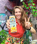 The image shows a woman smiling and holding a "Drinkin' Buds Beverages" Mai Tai can, while flashing a peace sign. She stands in front of a colorful graffiti wall with lush greenery, creating a fun and vibrant atmosphere.