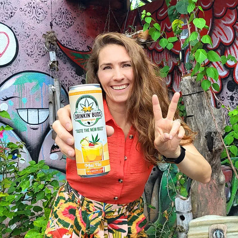 The image shows a woman smiling and holding a "Drinkin' Buds Beverages" Mai Tai can, while flashing a peace sign. She stands in front of a colorful graffiti wall with lush greenery, creating a fun and vibrant atmosphere.