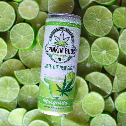 The image features a "Drinkin' Buds Beverages" Margarita can placed on a bed of fresh lime slices. The label highlights a cannabis leaf, the slogan "Taste the New Buzz," and a margarita graphic, emphasizing a cool, citrus-infused experience.