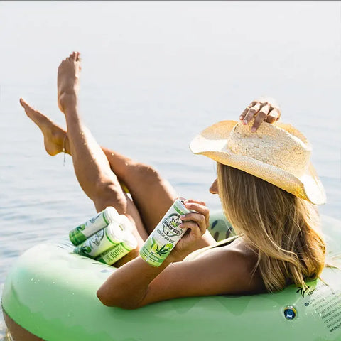 The image shows a woman lounging on an inflatable raft in a lake, wearing a straw hat and holding a "Drinkin' Buds Beverages" Margarita can. The scene conveys a relaxed, carefree vibe, perfect for enjoying a refreshing cannabis-infused drink outdoors.