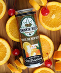 The image features a can of "Drinkin' Buds Beverages" in Wisconsin Old Fashioned flavor, lying on a wooden surface surrounded by fresh orange slices, orange peels, and bright red maraschino cherries. The arrangement emphasizes the drink's cocktail inspiration, highlighting its vibrant and refreshing essence.