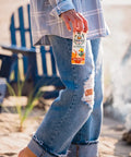 The image shows a person in ripped jeans and a plaid shirt holding a "Drinkin' Buds Beverages" Rum Punch can at a beach, with blue Adirondack chairs and a campfire in the background, creating a laid-back, coastal vibe.