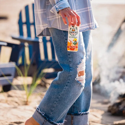 The image shows a person in ripped jeans and a plaid shirt holding a "Drinkin' Buds Beverages" Rum Punch can at a beach, with blue Adirondack chairs and a campfire in the background, creating a laid-back, coastal vibe.