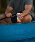 The image shows a person holding a "Drinkin' Buds Beverages" Rum Punch can while seated in a blue canoe, paddle in hand. The scene captures a relaxed, outdoor adventure vibe on the water.