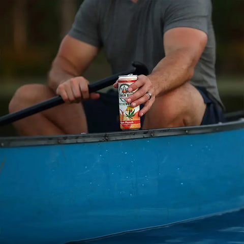 The image shows a person holding a "Drinkin' Buds Beverages" Rum Punch can while seated in a blue canoe, paddle in hand. The scene captures a relaxed, outdoor adventure vibe on the water.