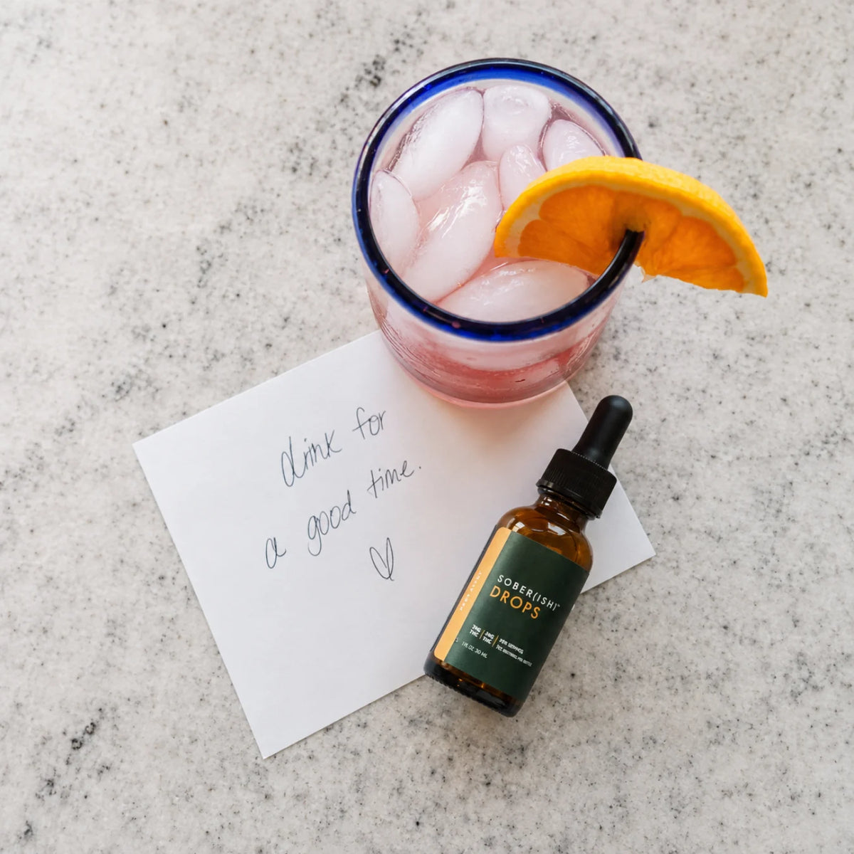 A pink drink with an orange slice next to a bottle of Sober(ish) Drops
